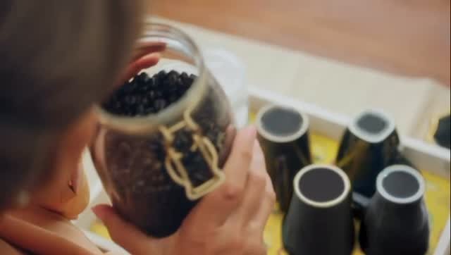 A person holding a glass jar filled with dark berries, with dark jars lined up on a wooden surface in front.